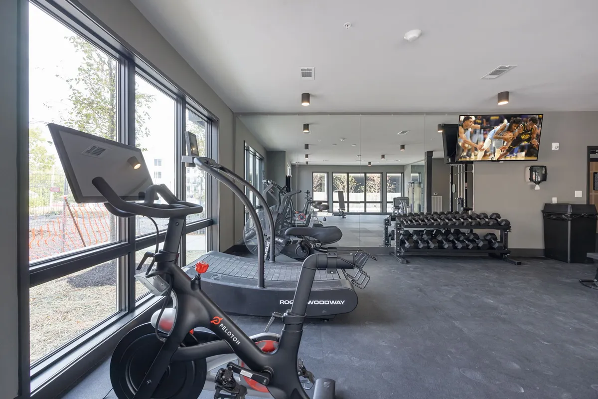 A bright fitness center equipped with a Peloton bike, treadmill, dumbbells, large windows, and a TV display. The space is modern, energetic, and flooded with natural light, inviting both solo workouts and group fitness classes.