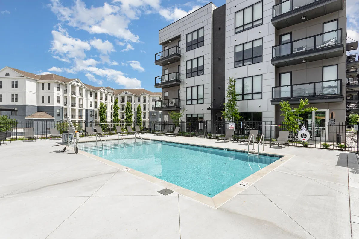 A lively scene featuring a sparkling swimming pool surrounded by landscaped greenery and balconies of modern apartments. The area is set up for social interactions, sunbathing, and relaxation, suggesting a vibrant communal atmosphere.