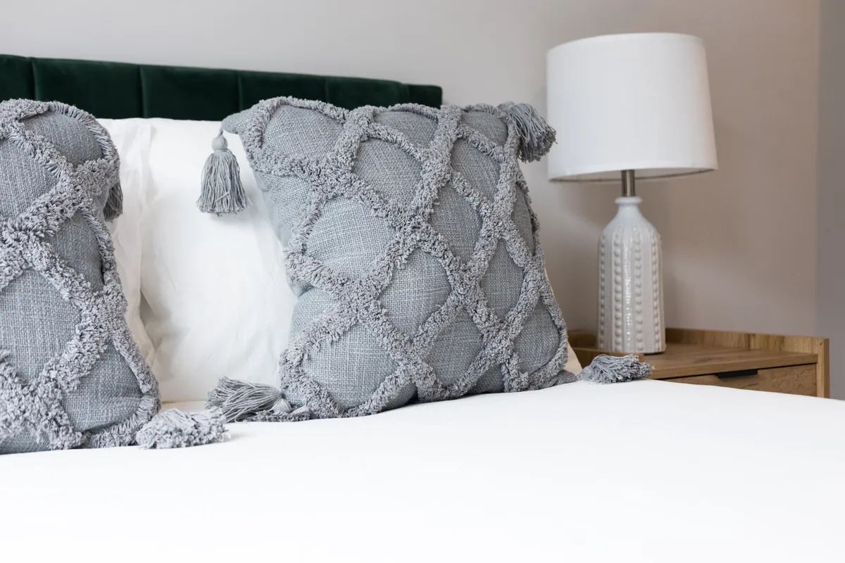 A cozy bedroom setup with decorative pillows, a bedside lamp casting a warm glow, and a nightstand. The decor is inviting, suggesting restful sleep and moments of relaxation.