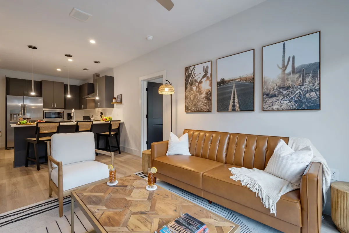 The hero image showcases a sunlit, inviting living space with a plush leather sofa, a sleek wooden coffee table, stylish framed wall art, and an open kitchen bar area perfect for gatherings. The decor blends modern simplicity with cozy accents, highlighting the home’s warm and welcoming vibe.