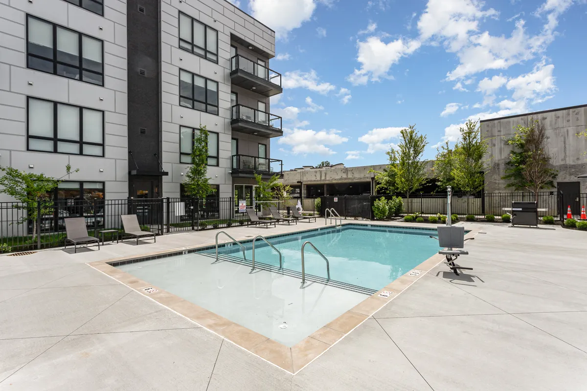 An exterior view of a swimming pool adjacent to the apartment building, surrounded by landscaped greenery and a balcony. The setting is ideal for social gatherings, sunbathing, or barbecues, with a sense of community and relaxation.