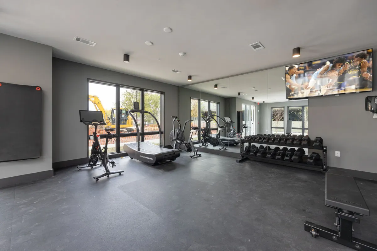 A bright, modern fitness center featuring a treadmill, weight rack, exercise bike, and television. The space is designed for energetic workouts and group fitness, with natural light and an inviting atmosphere.