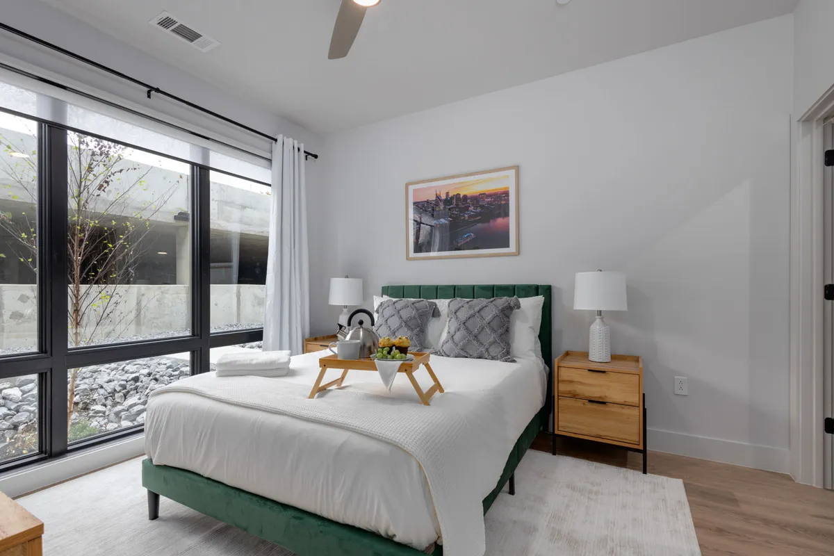 A serene bedroom featuring a green upholstered bed, large windows allowing for ample natural light, decorative artwork, and bedside tables. The overall atmosphere is peaceful and inviting, ideal for relaxation and restful sleep.