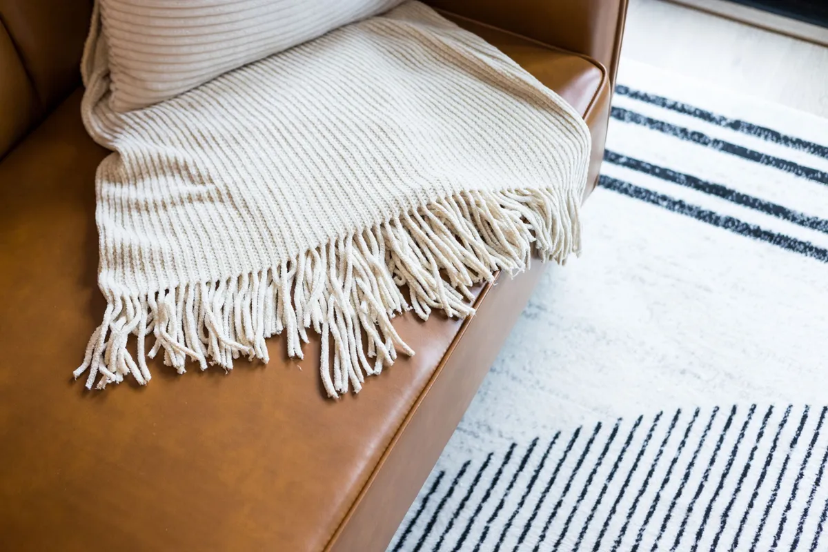 A warm, inviting living room area with a brown leather sofa, a knitted blanket draped over the arm, and a striped rug underfoot. The room feels homey and perfect for families to spend time together.