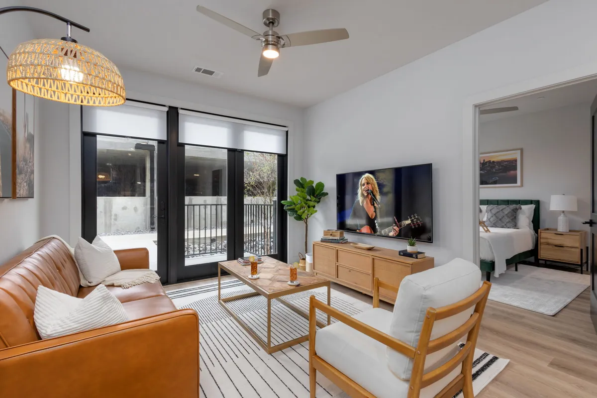 A stylish living room with a leather sofa, a TV showing a live performance, a wooden coffee table, and lush indoor plants. The room is bright, comfortable, and inviting, perfect for relaxing or gathering as a group.
