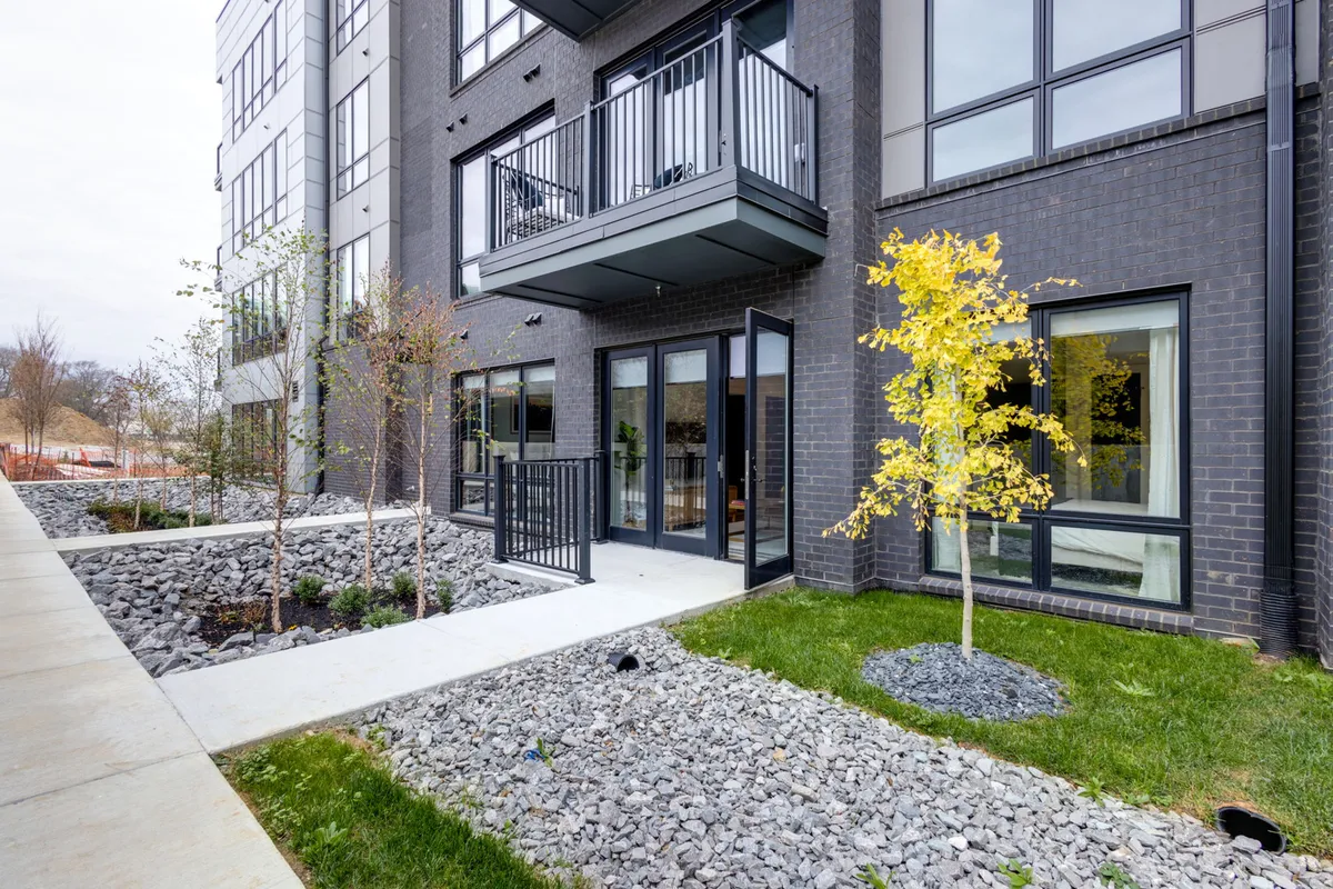 A modern balcony with a yellow-leaved tree and decorative gravel sits outside large glass doors, hinting at the private patio spaces available. The setting is contemporary and inviting, ideal for relaxing or taking work outside.
