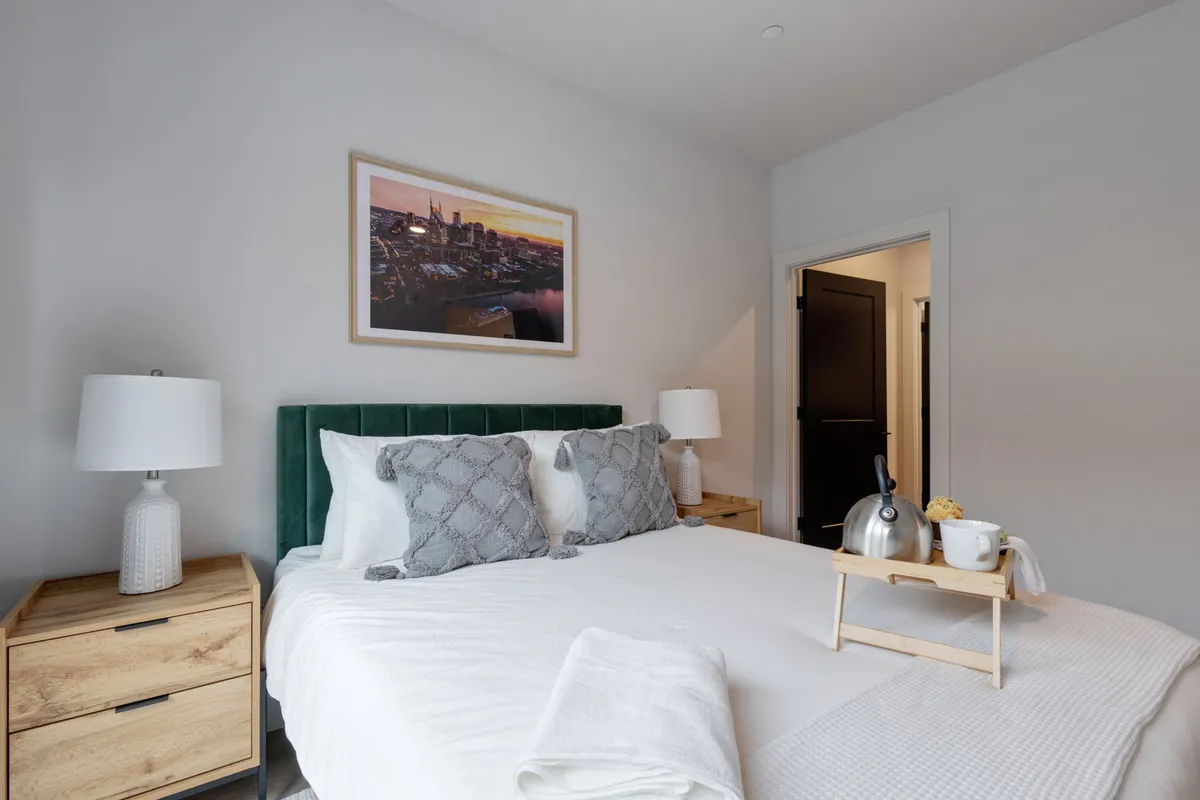 A tranquil bedroom with a green upholstered headboard, framed cityscape artwork, a breakfast tray with kettle and cups, and nightstands with lamps. The room exudes comfort and a sense of waking up refreshed, with thoughtful touches for morning routines.